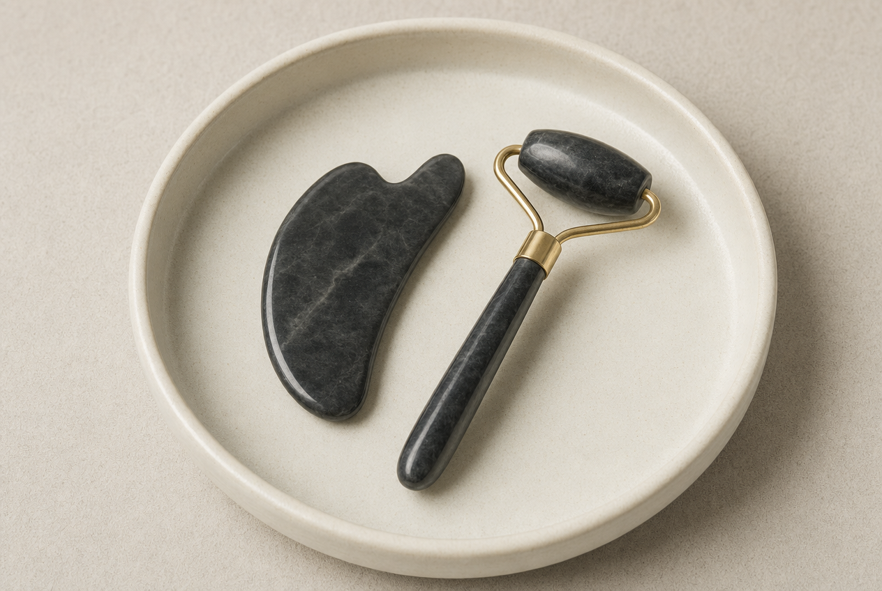 A dark marbled stone tool and a matching facial roller with brass fittings resting together in a shallow cream ceramic dish, representing the two techniques compared in gua sha research, Belldiva